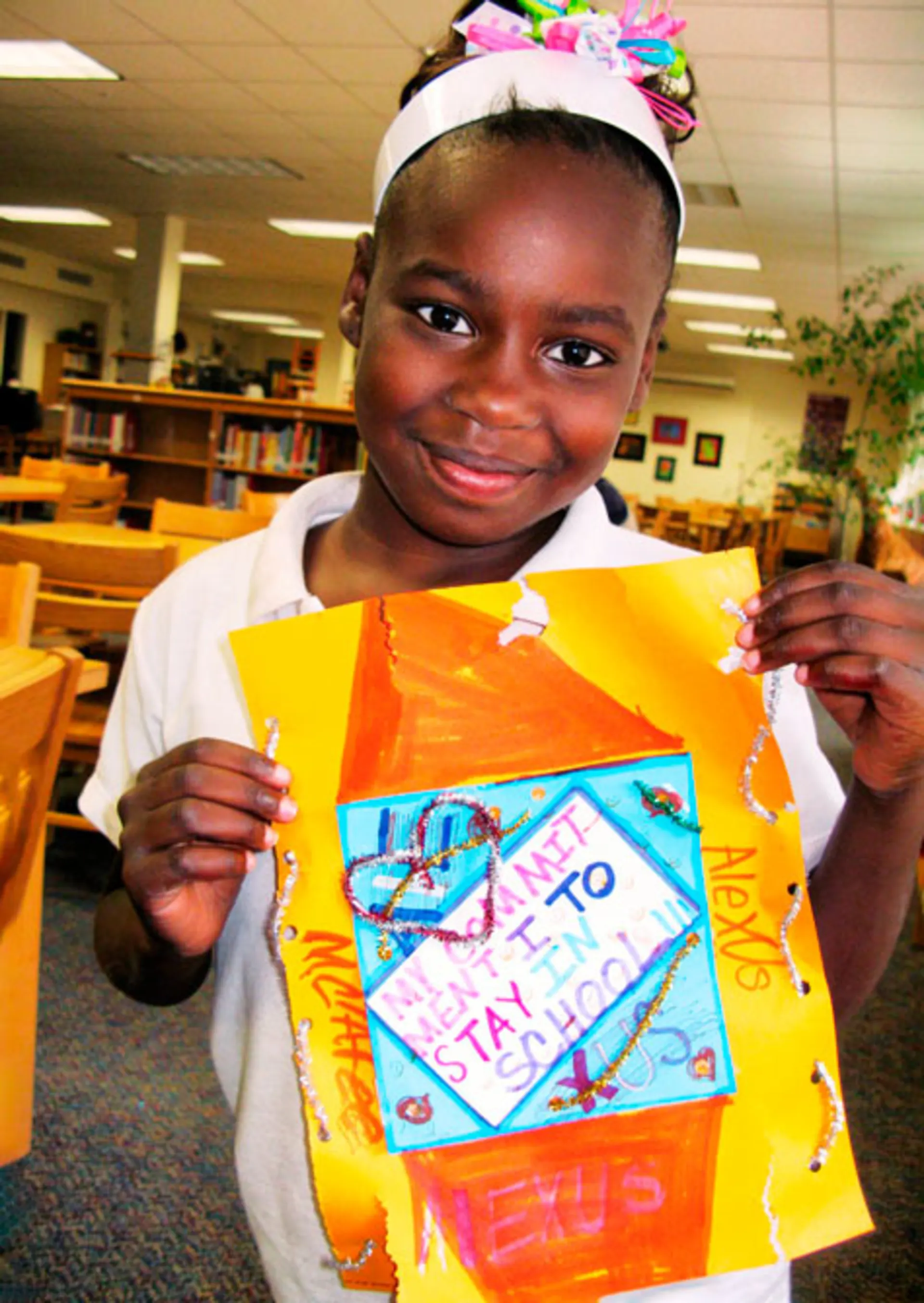 A different young girl standing in a library smiles and holds up a collage with the text 'My commitment is to stay in school.'.