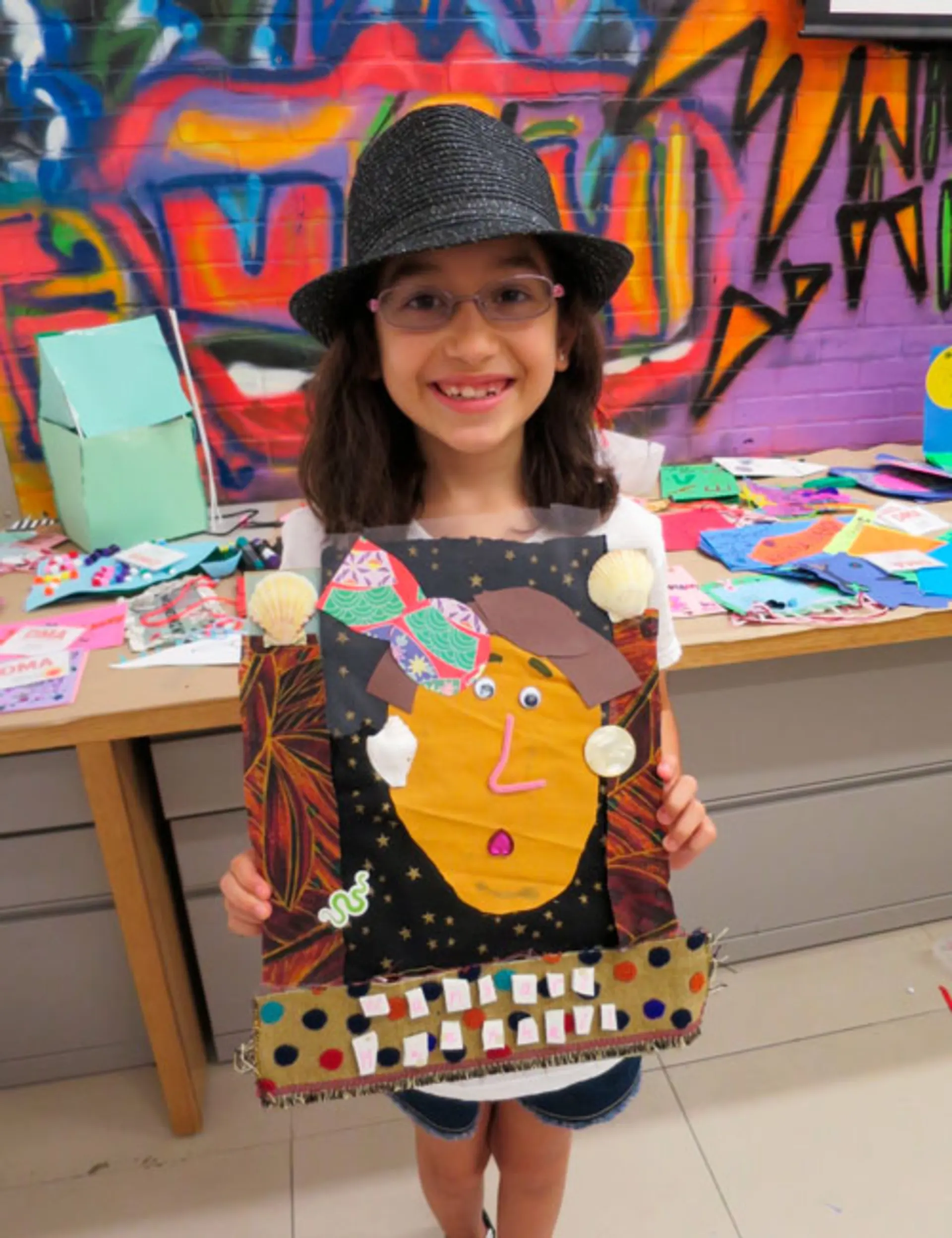 A young girl standing in front of a table of art supplies smiles and holds up a collage portrait of a peacemaker.