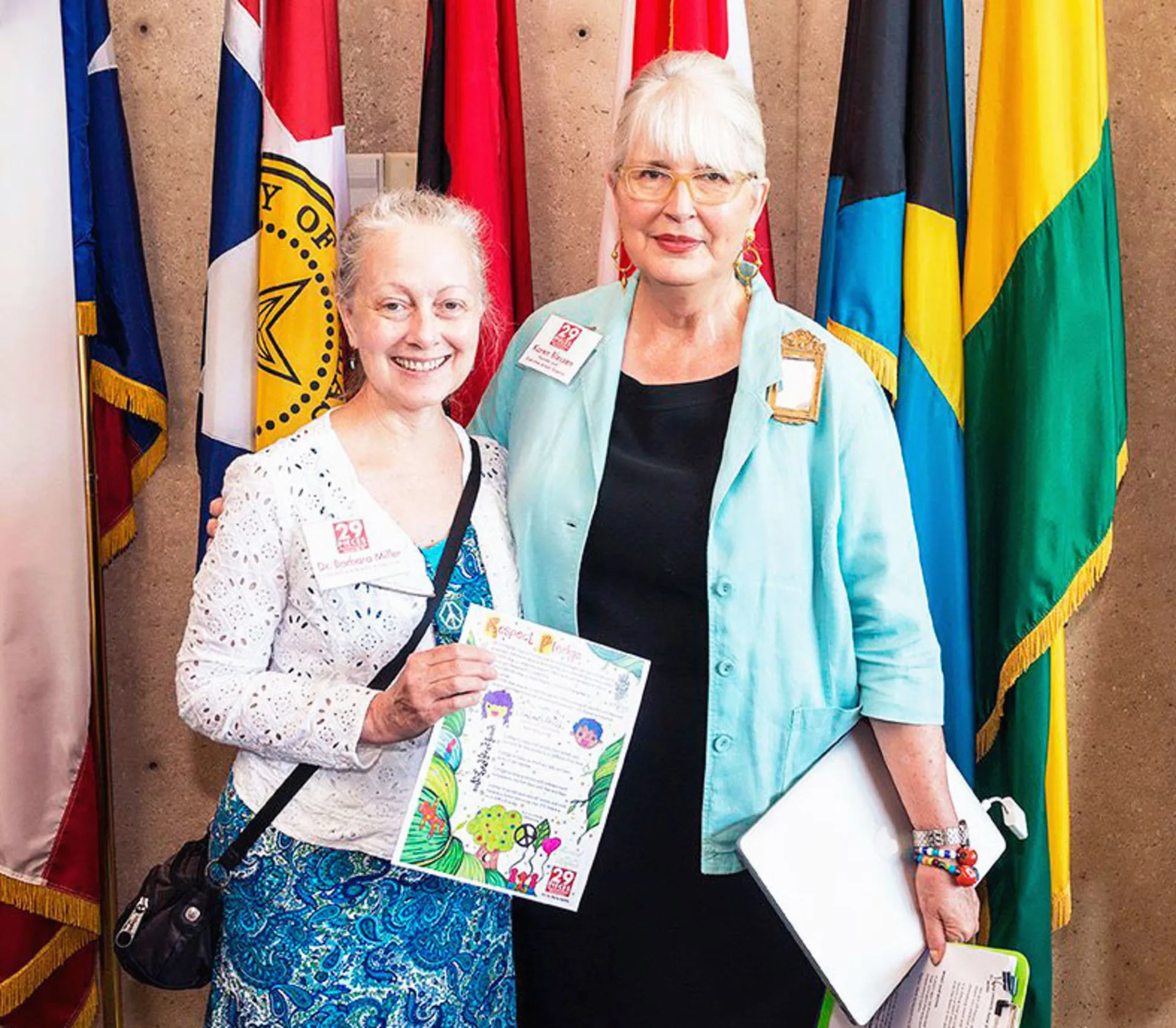 Blessen and Dr. Miller, who are both holding copies of the Respect Pledge, which was used in 29 Pieces' 2016 Dallas Respect Project, smile while standing in front of a row of flags.