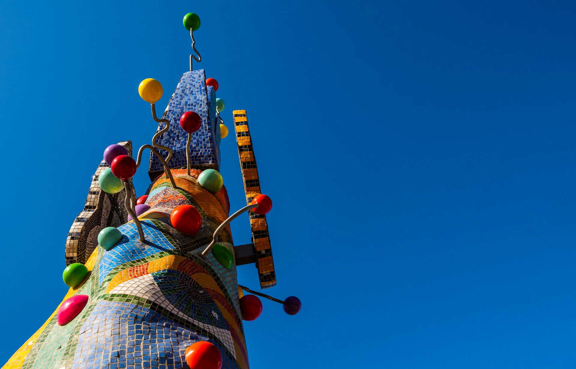 A low-angle view of the colorful, abstract Piece 24 sculpture set against a deep blue, cloudless sky. The base of the sculpture is adorned in bright, multicolored spheres and mosaic tiles that form patterns, including a large human eye.