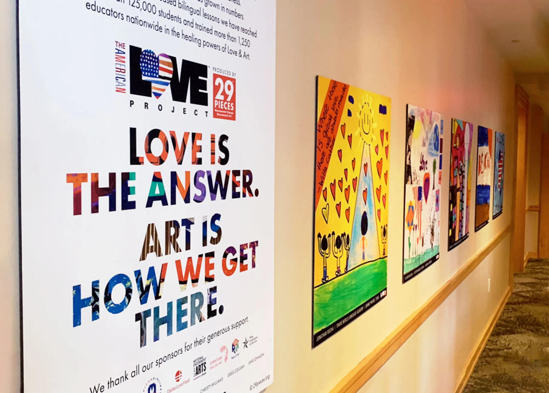 Hallway display with five large mounted prints of student artwork. In the foreground is a sign with The American Love Project logo and the text, “Love is the answer. Art is how we get there.”