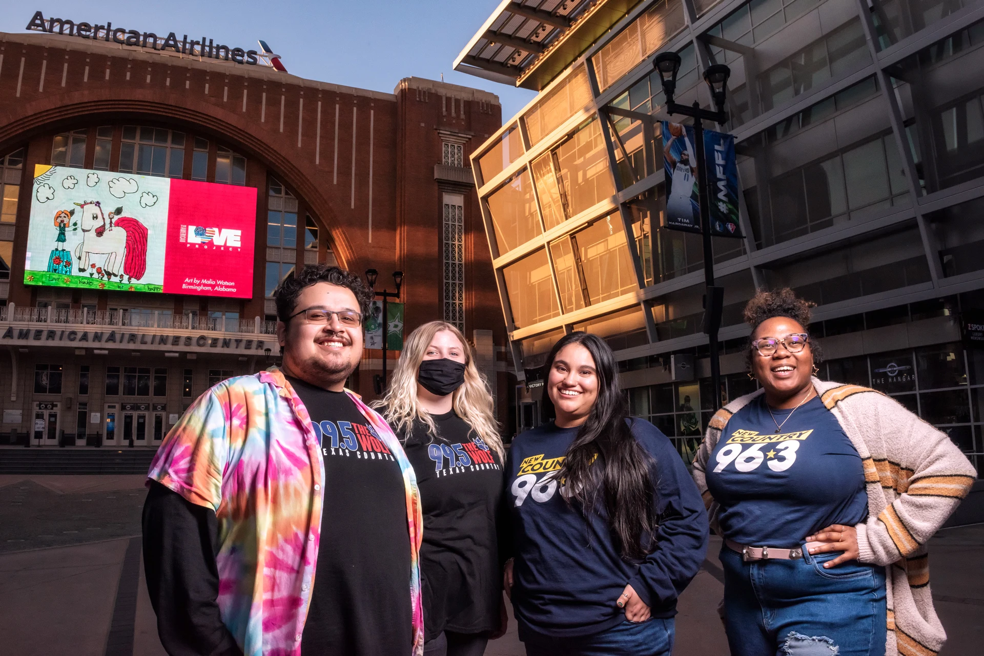 A group of friends smile and pose outside the plaza entrance of the American Airlines Center. Behind them on the digital display is a childlike drawing of a girl feeding apples to a unicorn. To the right is The American Love Project logo and the text “Art by Malia Watson, Birmingham, Alabama”