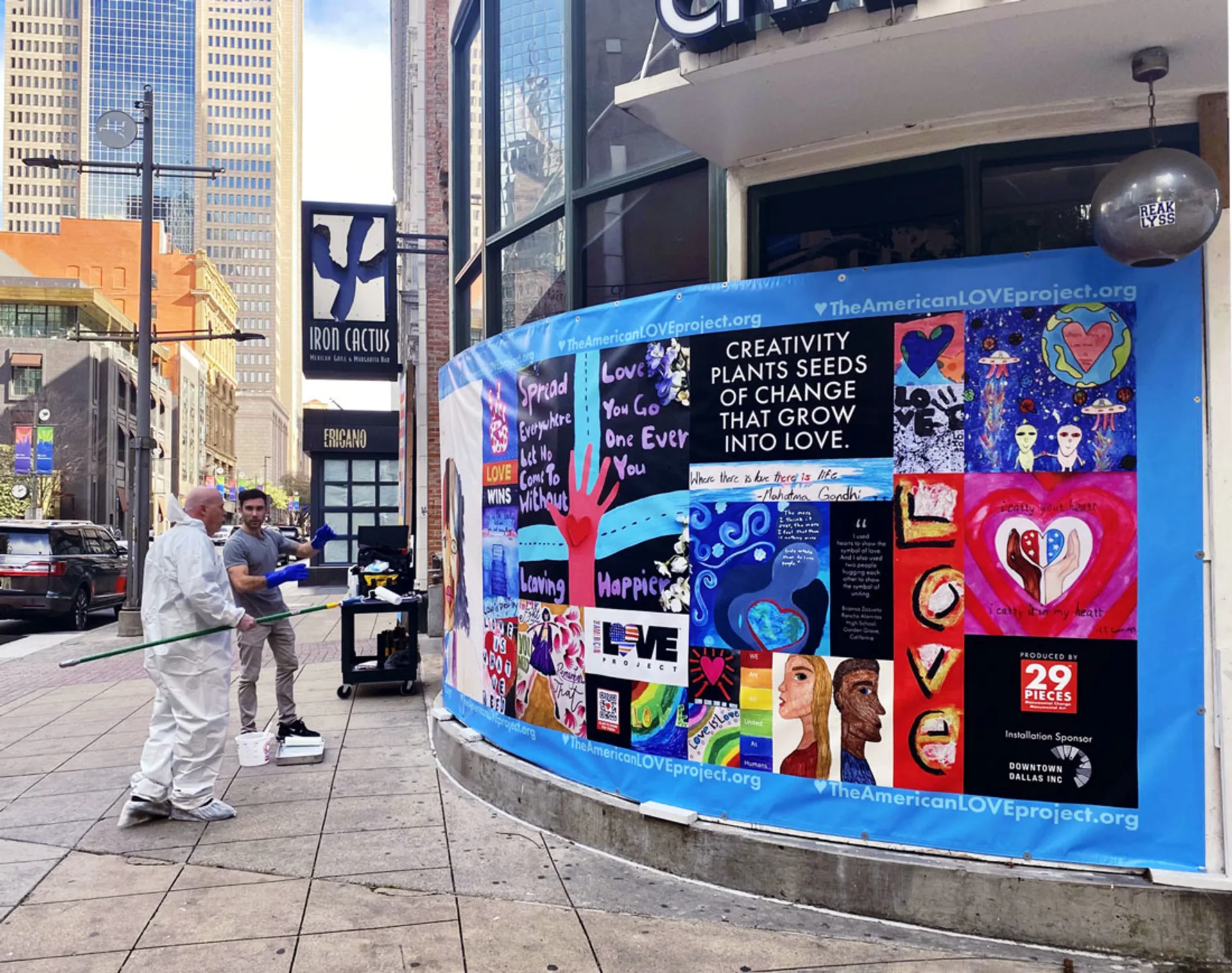 Two men install a large banner of colorful American Love Project artwork, which is interwoven with messages about love and creativity, along the curved exterior of the Iron Cactus restaurant on a street corner in downtown Dallas.