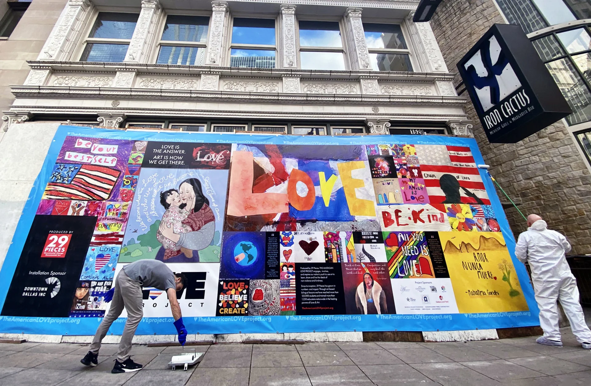 Street view of two men rolling adhesive over a large colorful banner installation featuring student art near the Iron Cactus restaurant sign. In the bottom left corner of the banner is the text “Produced by 29 Pieces,” and “Installation Sponsor: Downtown Dallas Inc.”