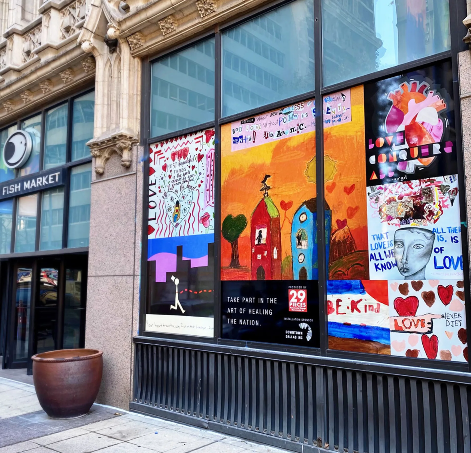Street-level window display featuring colorful student artwork outside a restaurant in downtown Dallas. In the center of the installation is the text, “Take part in the art of healing the nation,” next to the 29 Pieces logo and the Downtown Dallas Inc. sponsor logo. 