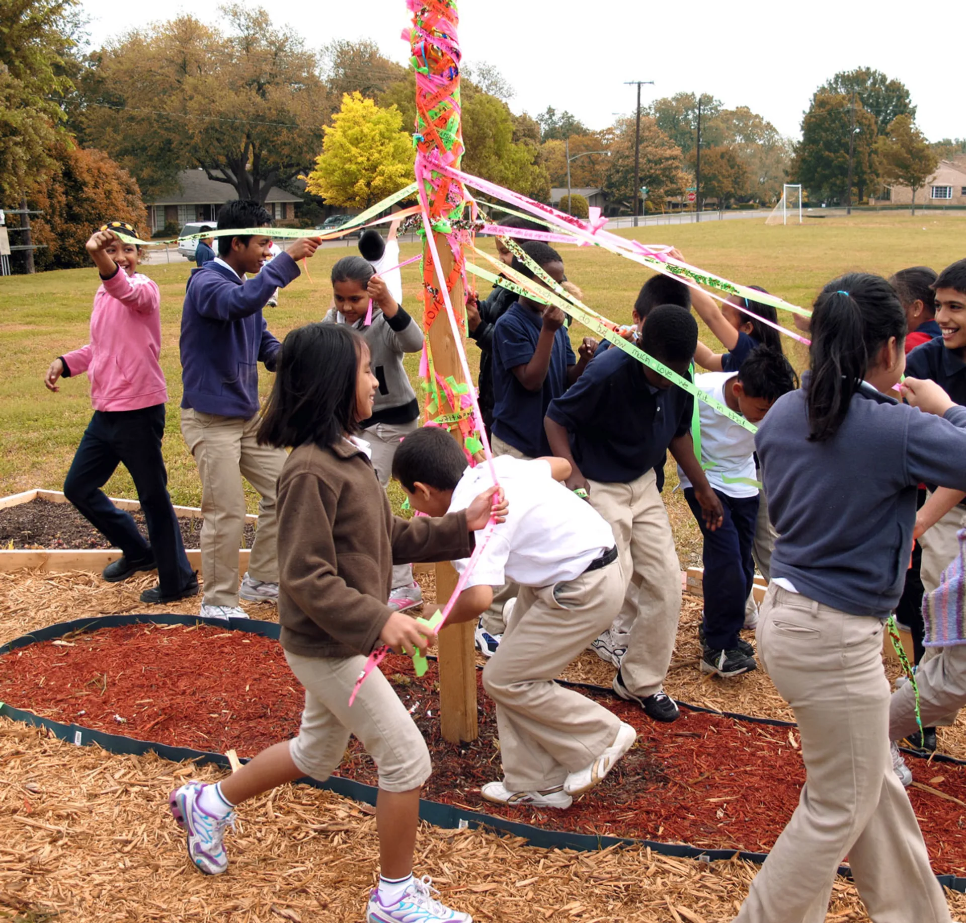 A group of middle school students cover a wooden pole in a park with layers of pink, orange and green ribbon by running around the pole in circles while holding onto the end of each ribbon.