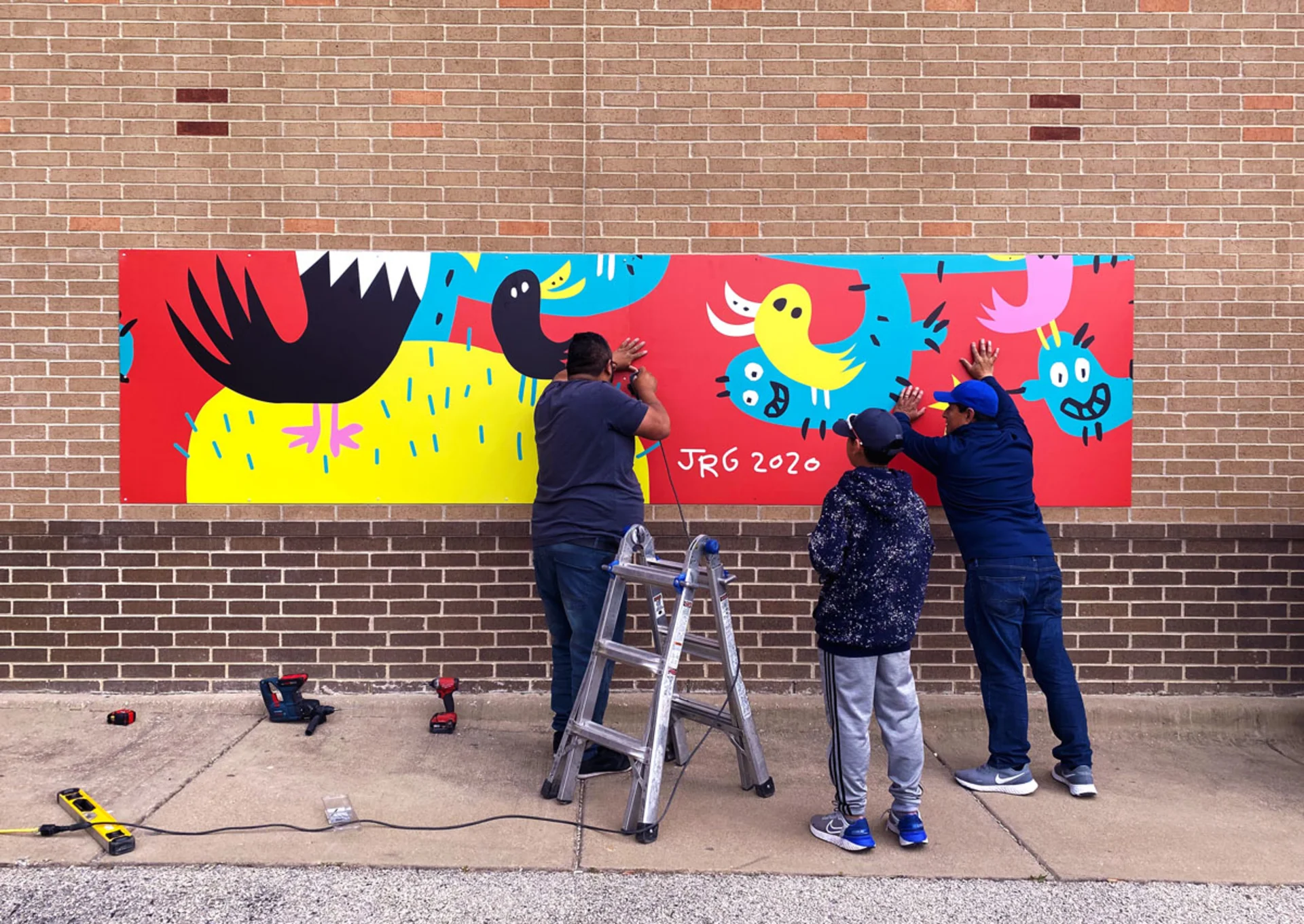 A member of the Thomas Printworks crew uses a screw to install a section of the Salazar Elementary mural along the school's brick exterior while other workers help.
