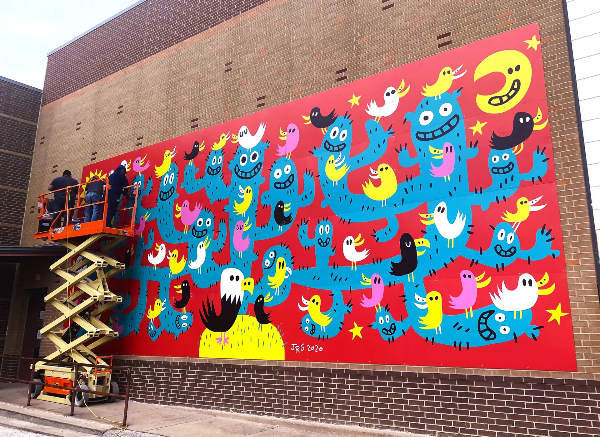 Thomas Printworks crew members on a lift finishing the installation of the Salazar Elementary mural, which depicts smiling blue cacti, birds, a moon and stars against a red background.
