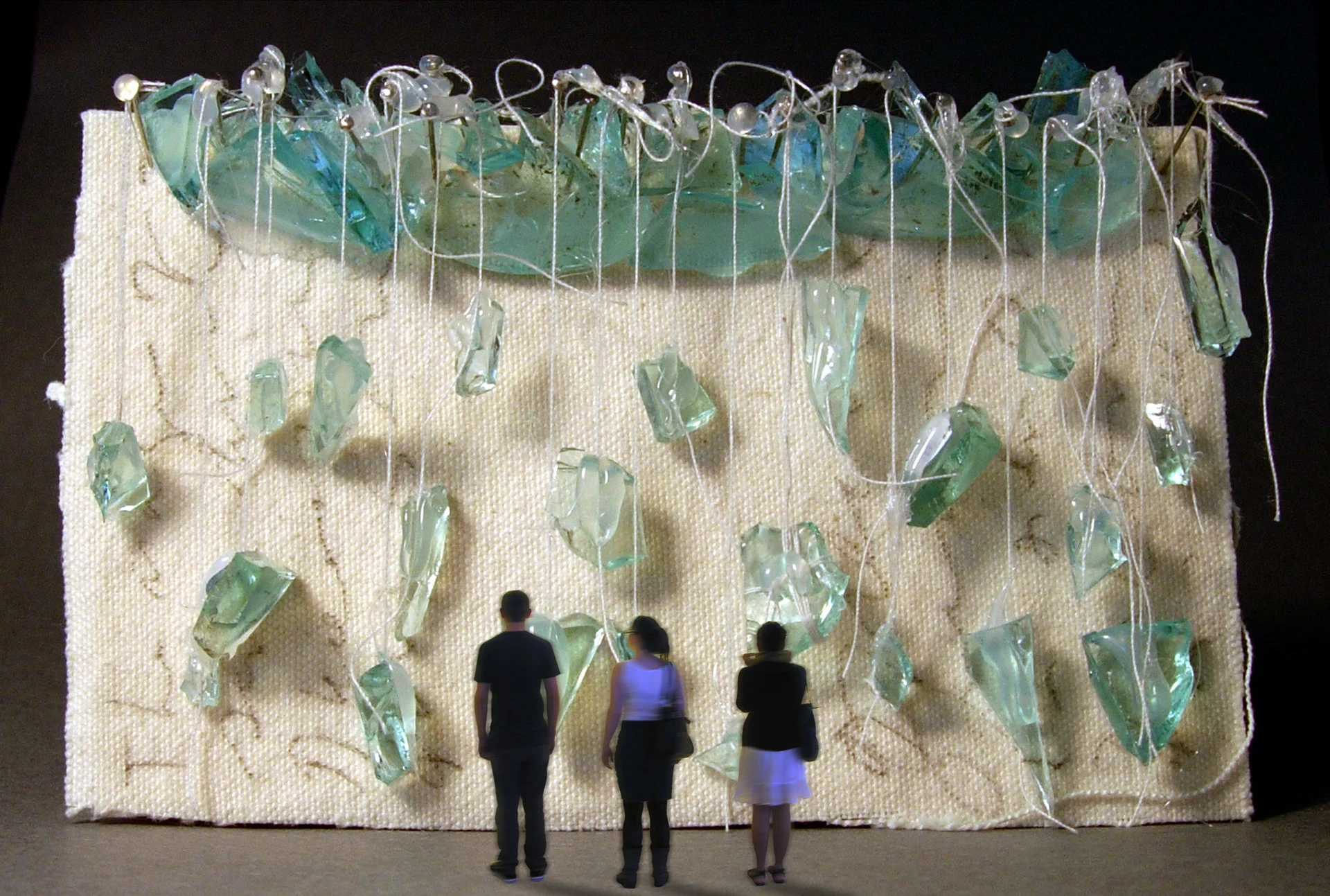 Visualization of a large beige woven structure decorated at the top with pieces of blue-green glass, which resemble sea glass, and giant sewing pins holding up pieces of string, from which more glass is hanging. Three blurred figures stand in the foreground looking at the art.