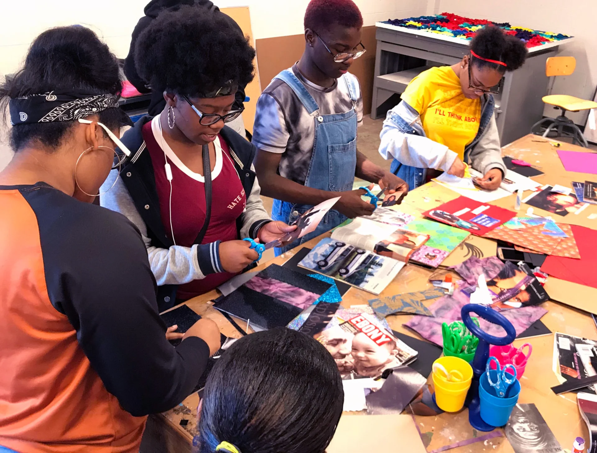 A group of students gather around a table and use scissors to cut sections out of magazines and multicolored pieces of paper for the collages they're making.