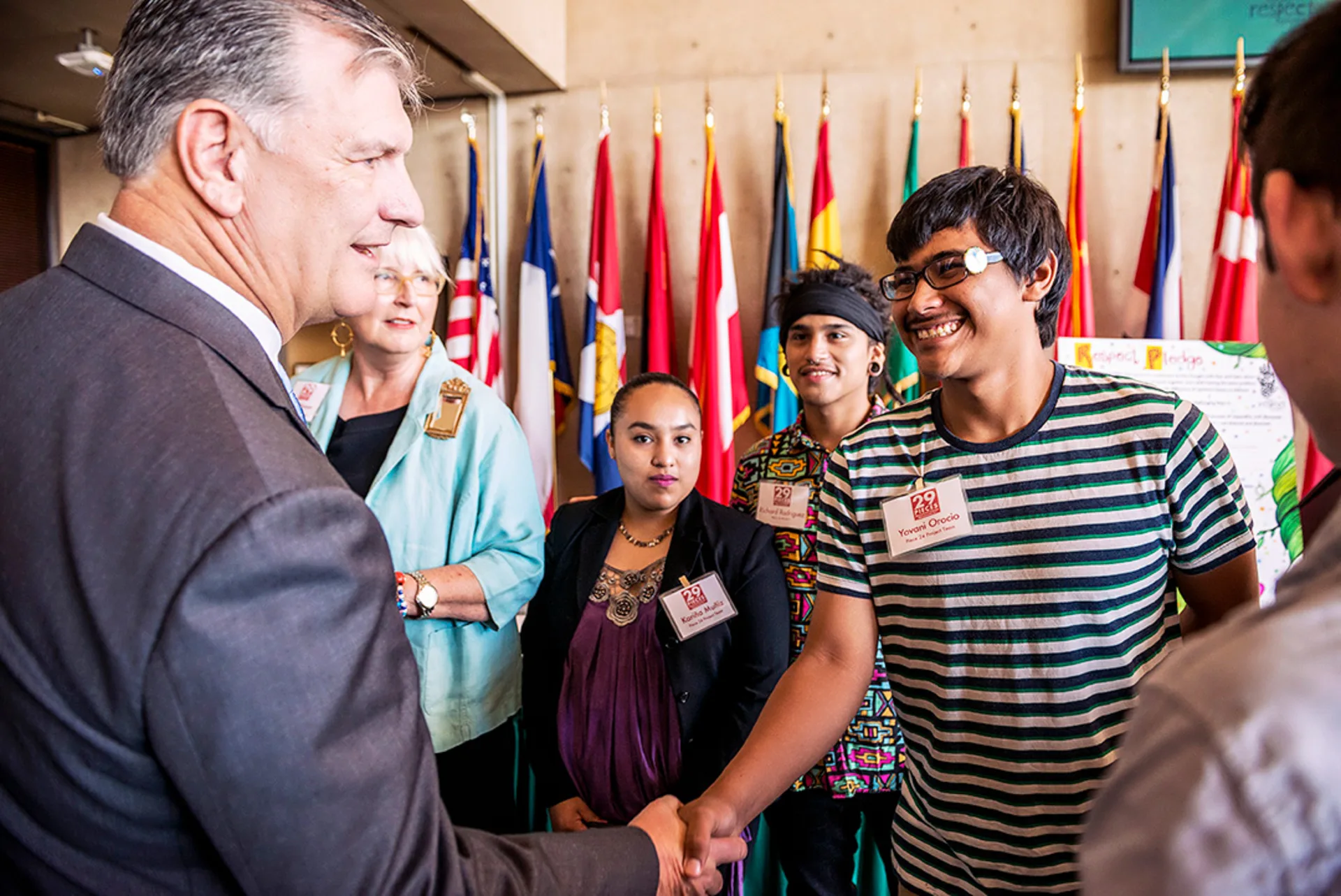 Karen Blessen and a group of high school student interns stand in a brightly lit room with flags as the Dallas mayor leans over to shake the hand of Orocio, who is smiling.