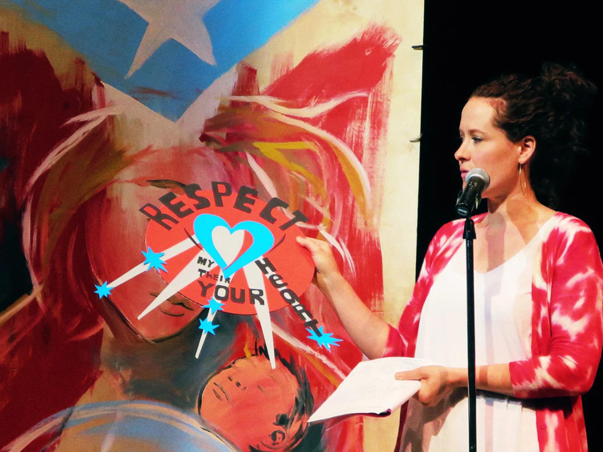 A teacher stands on stage near a microphone while holding a collage with rays extending from a heart and the text, 'Respect my, their, your heart'.