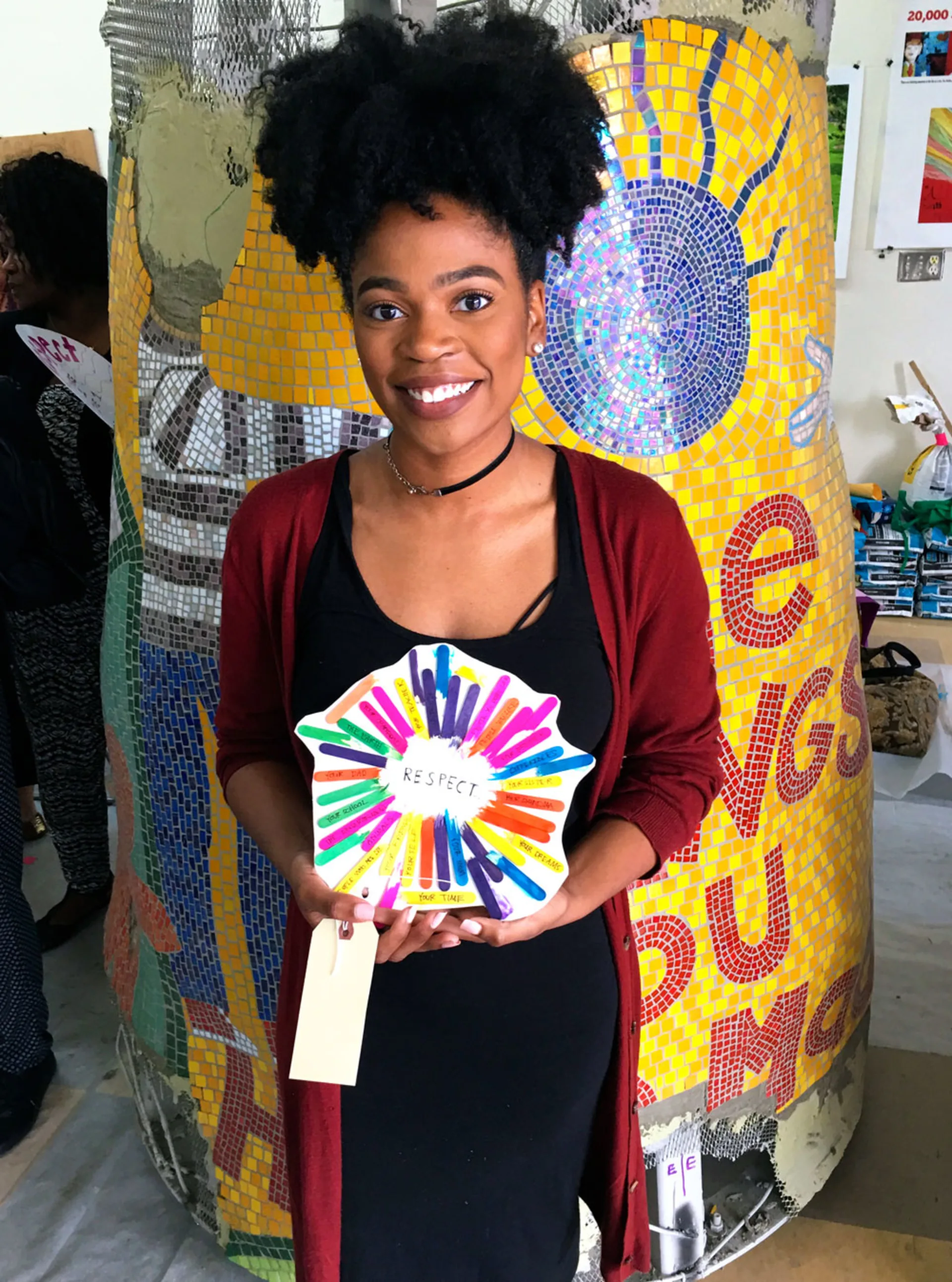 A woman smiles while standing in front of a mosaic sculpture and holding up a star shaped piece of art, which includes brightly colored popsicle sticks extending like rays from the word 'Respect'.