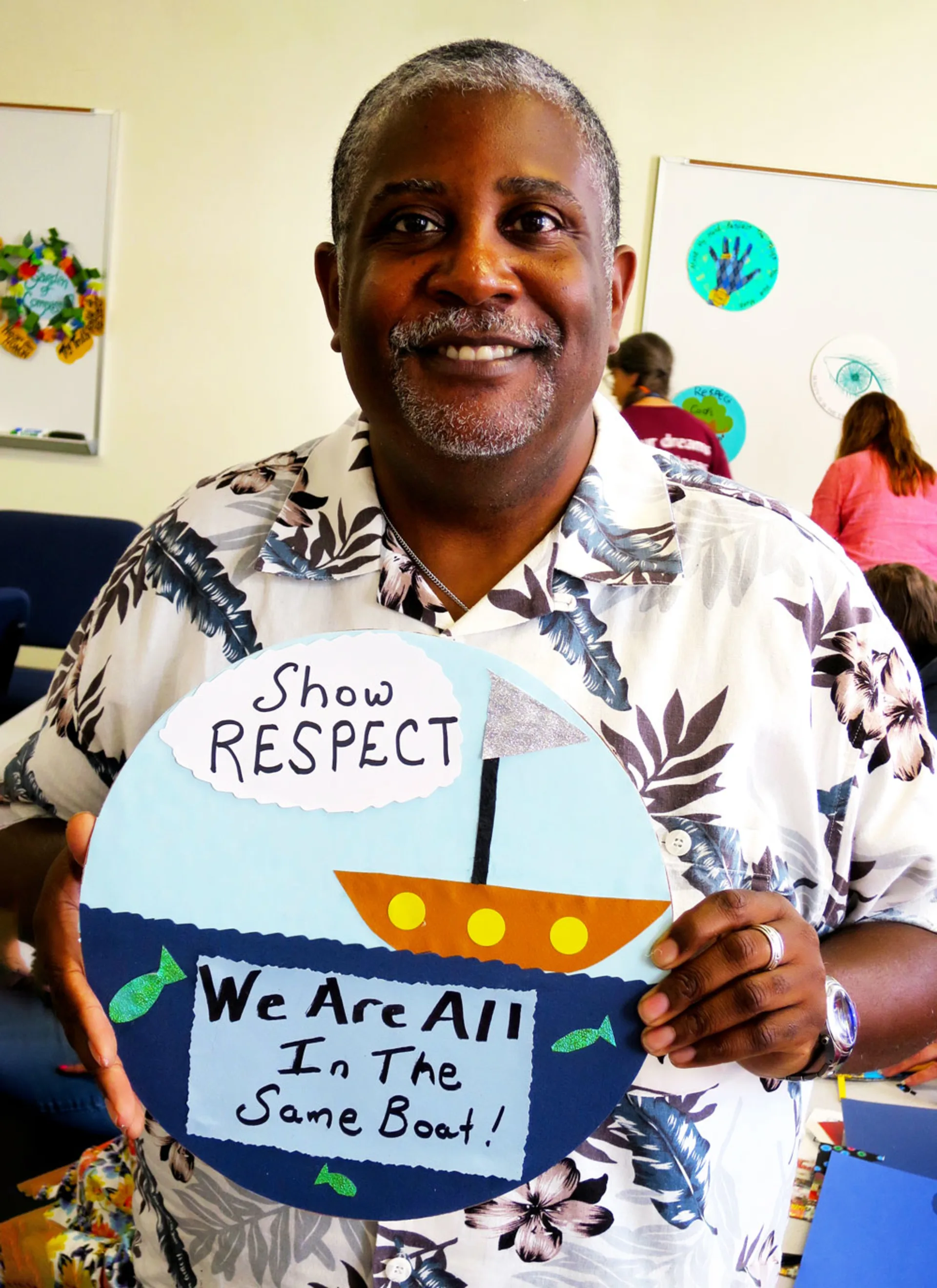 A smiling man wearing a Hawaiian shirt holds up a collage with a boat and underwater fish along with the text, 'Show respect. We are all in the same boat!'.