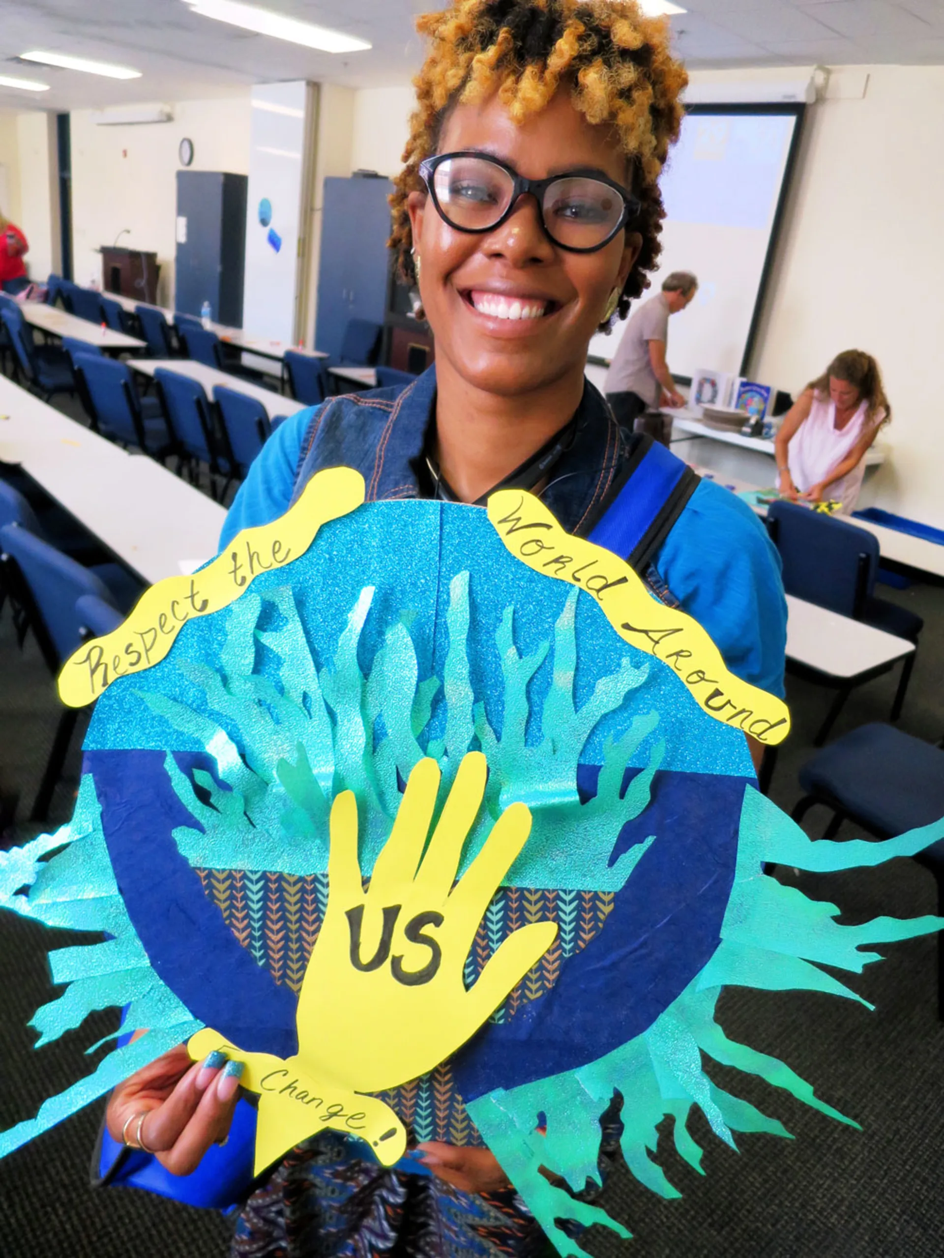 A smiling woman holds a colorful handmade collage with a yellow hand labeled 'Us' surrounded by turquoise paper shapes resembling ocean coral. A yellow ribbon above the hand reads, 'Respect the World Around Us'.