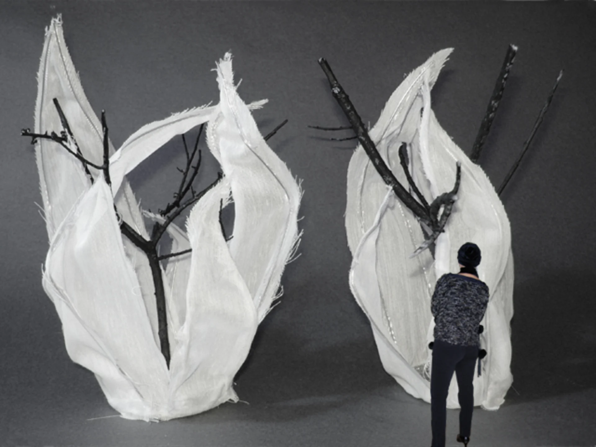 Pieces of white cloth resembling flames is draped around two tree-like twig structures in a gallery enveloped in gray walls and floor, while a woman looks at the art.