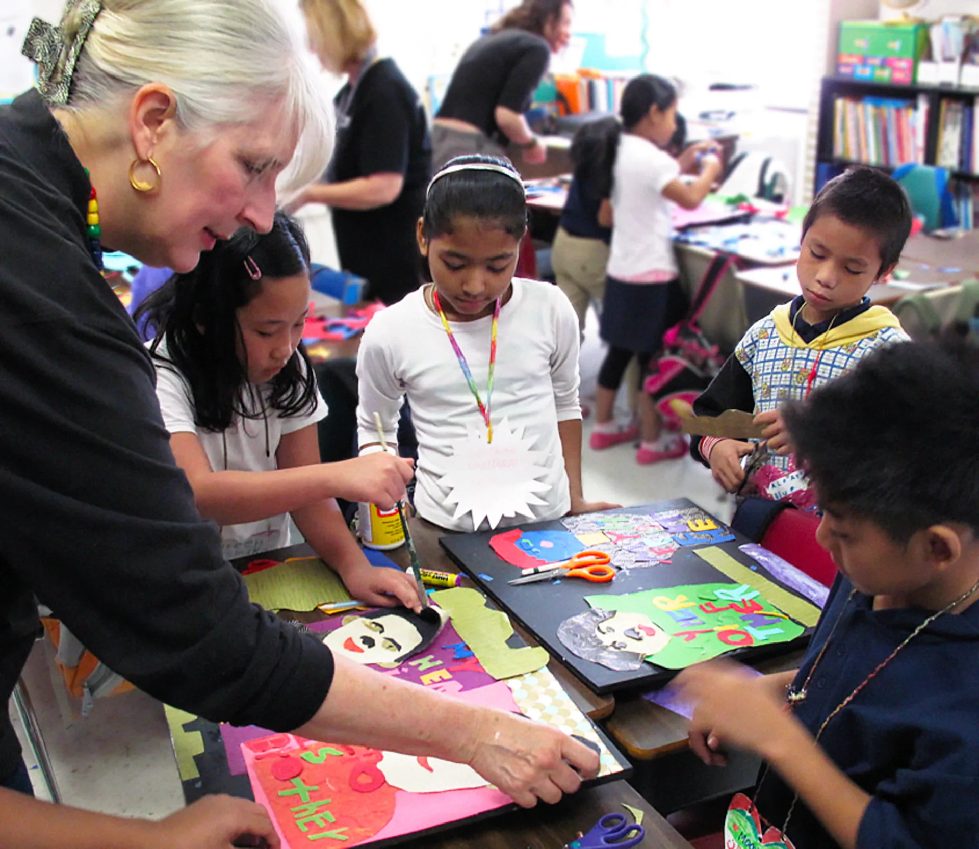 29 Piece co-founder Karen Blessen and a group of young children gather around a classroom table and work intently on a portrait collage art project using paintbrushes, scissors and glue.
