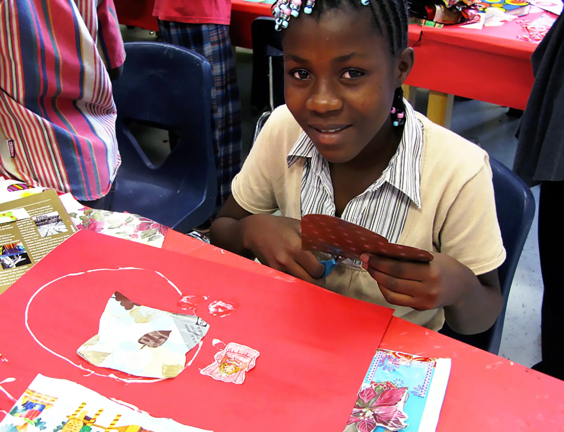 A young girl with beaded dreadlocks sits a table while cutting a heart out of a piece of paper for a collage.