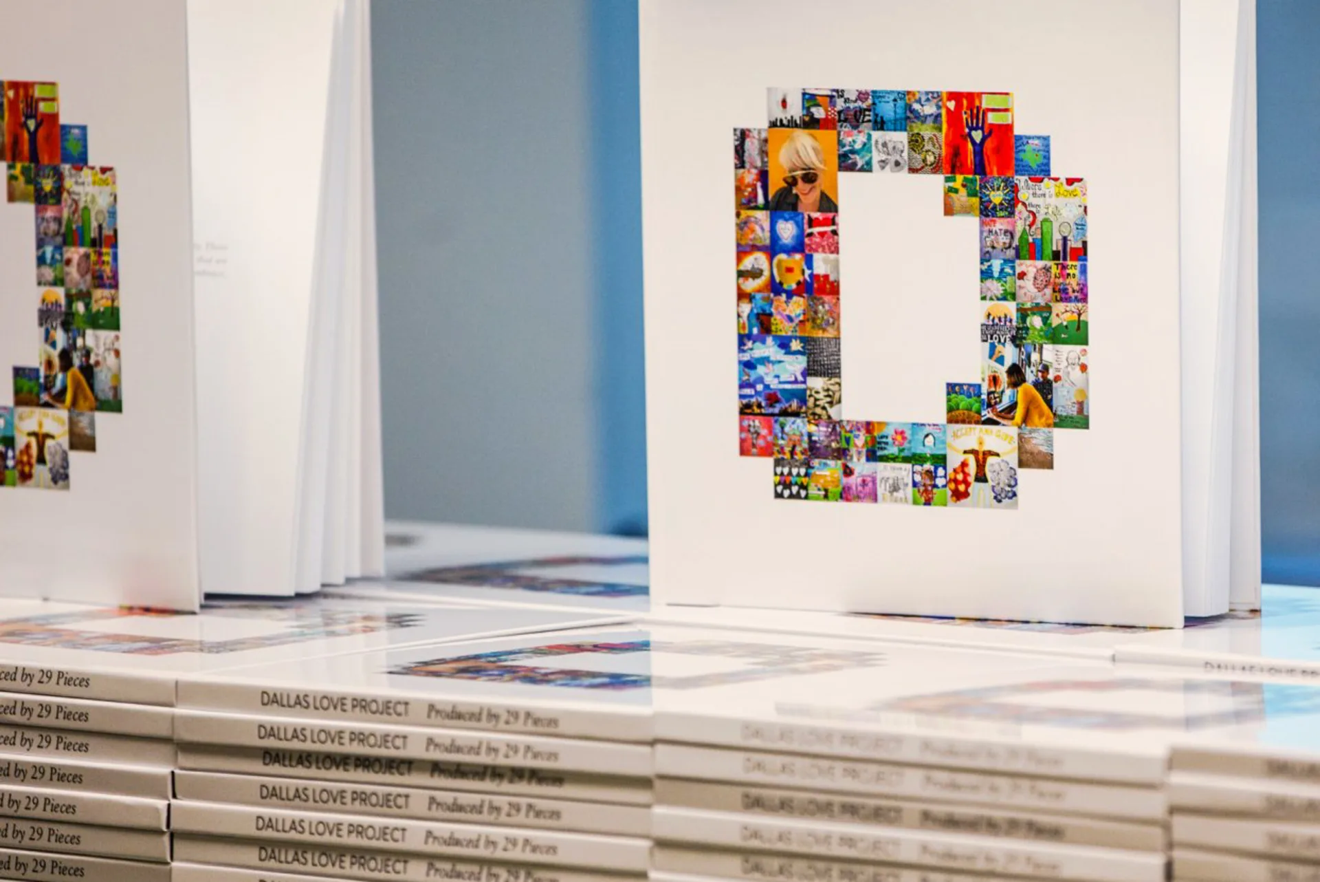 Rows of stacked white hardcover books with the words, ‘Dallas Love Project, Produced by 29 Pieces’ on each spine. On top of the stacks, several books are displayed; their front covers are illustrated with dozens of small photographs of people and art that form the letter D for Dallas.