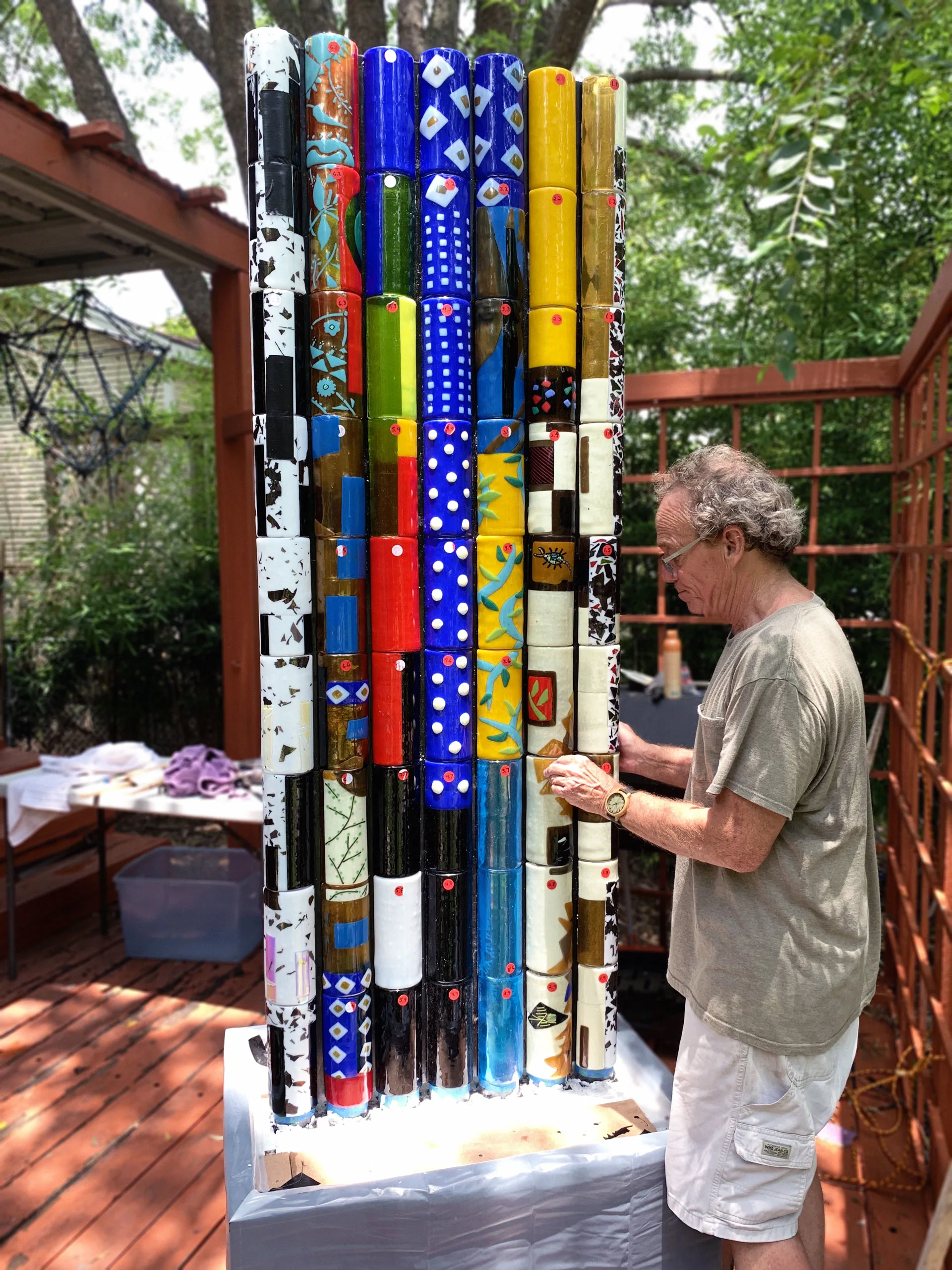 Joe Stokes looks down while holding part of the Piece 10 sculpture, which is constructed from tall, multicolored glass cylinders aligned in rows. Trees are in the background.