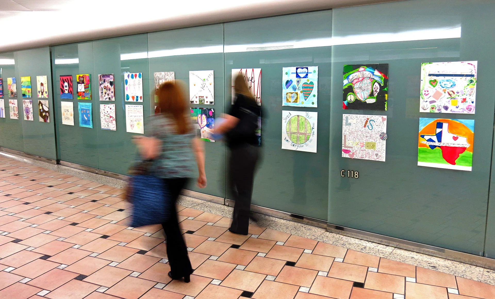 Two people walk past a wall displaying colorful children's artwork inside a building with tiled floors and frosted glass panels marked 'C 118'.