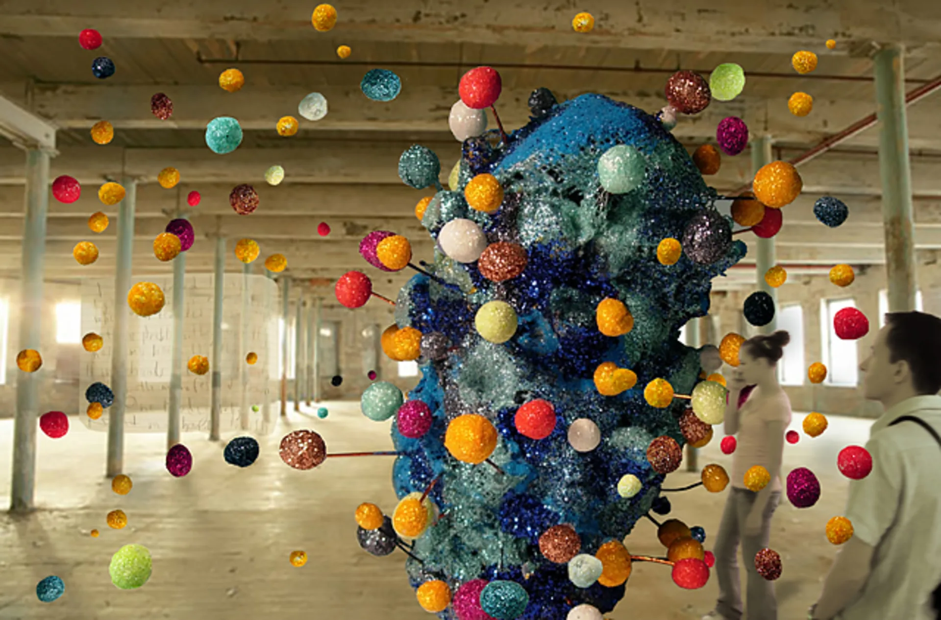 Colorful, glittery spheres on wires protrude from a large, abstract blue sculpture in an industrial warehouse, with two blurred figures walking in the background.