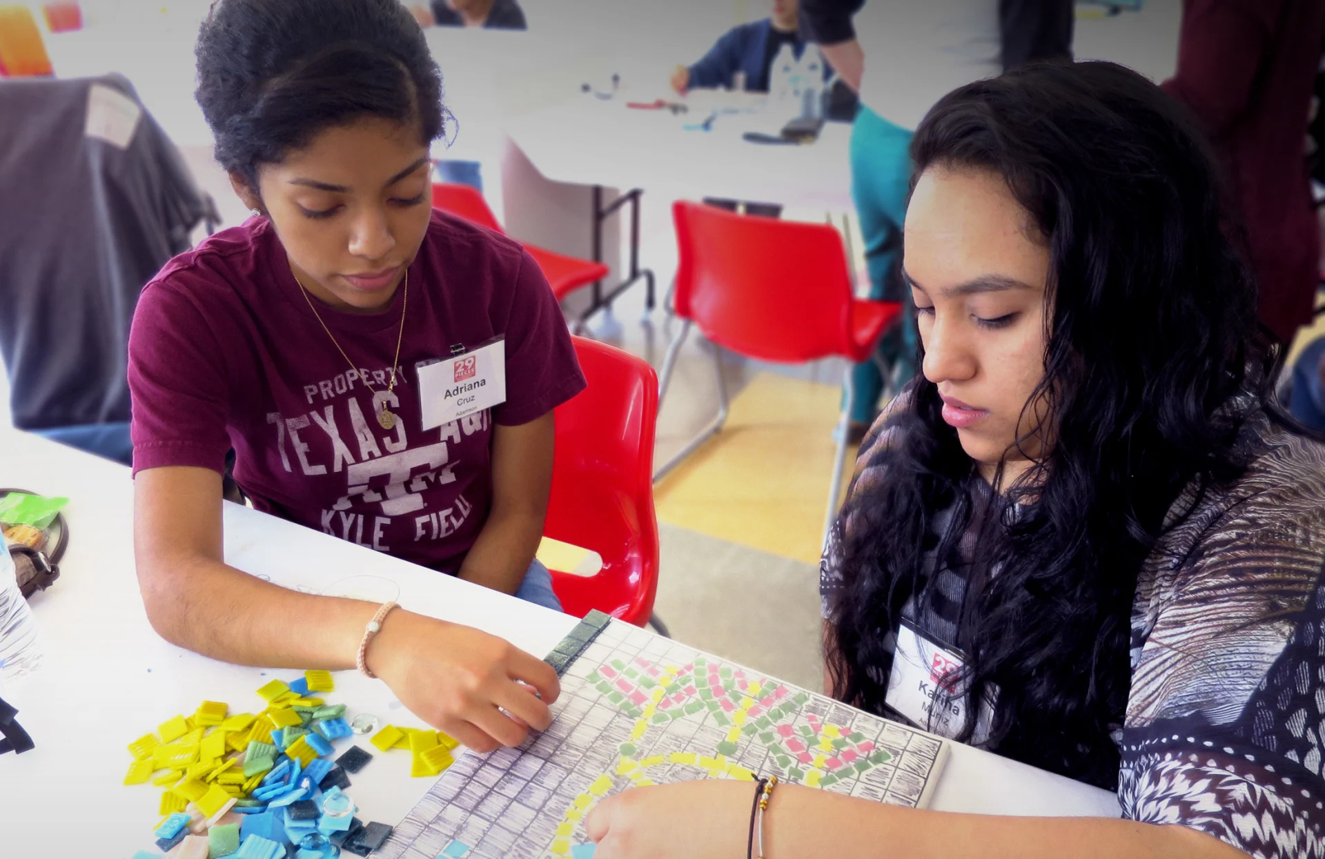 Two young women sitting at a table working intently together on a colorful tile mosaic, with small square tiles in different colors spread out in front of them.