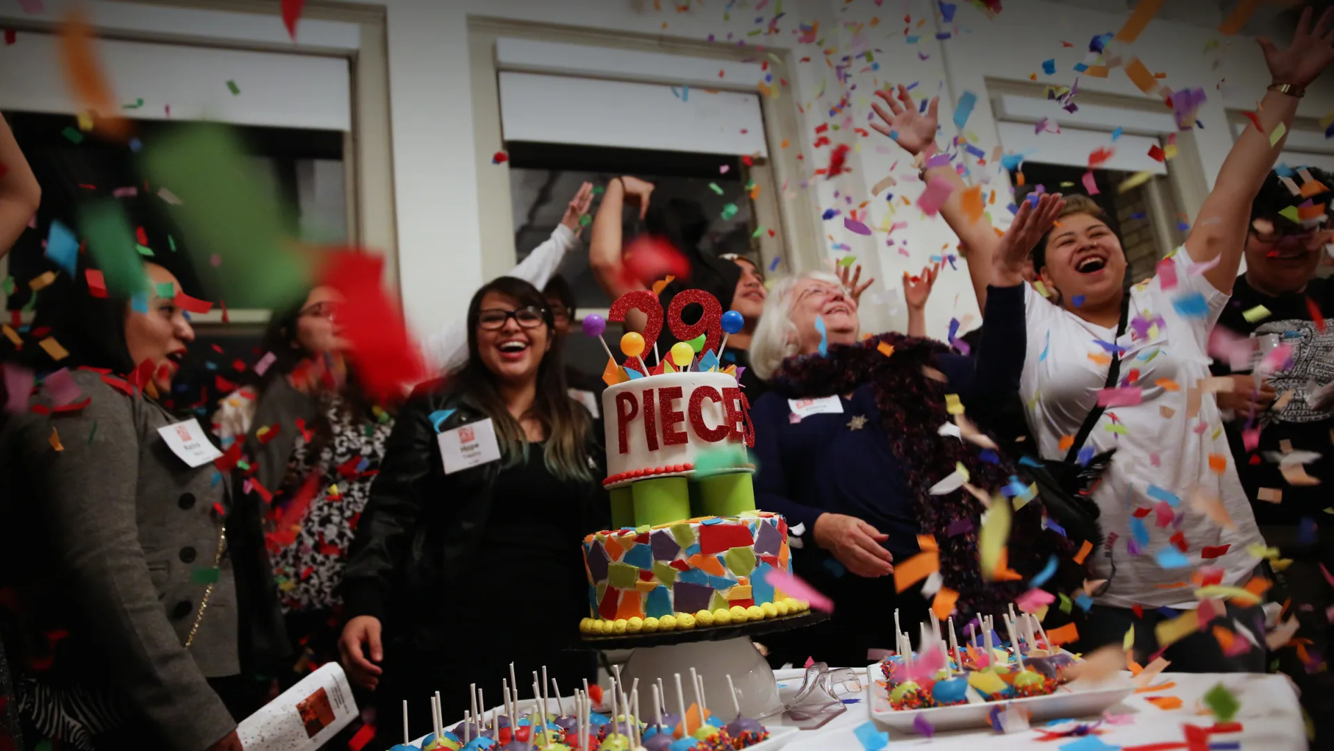 A large group of 29 Pieces members celebrate at the organization's 10 year anniversary party, smiling and cheering next to a cake with the words '29 Pieces' as confetti falls.
