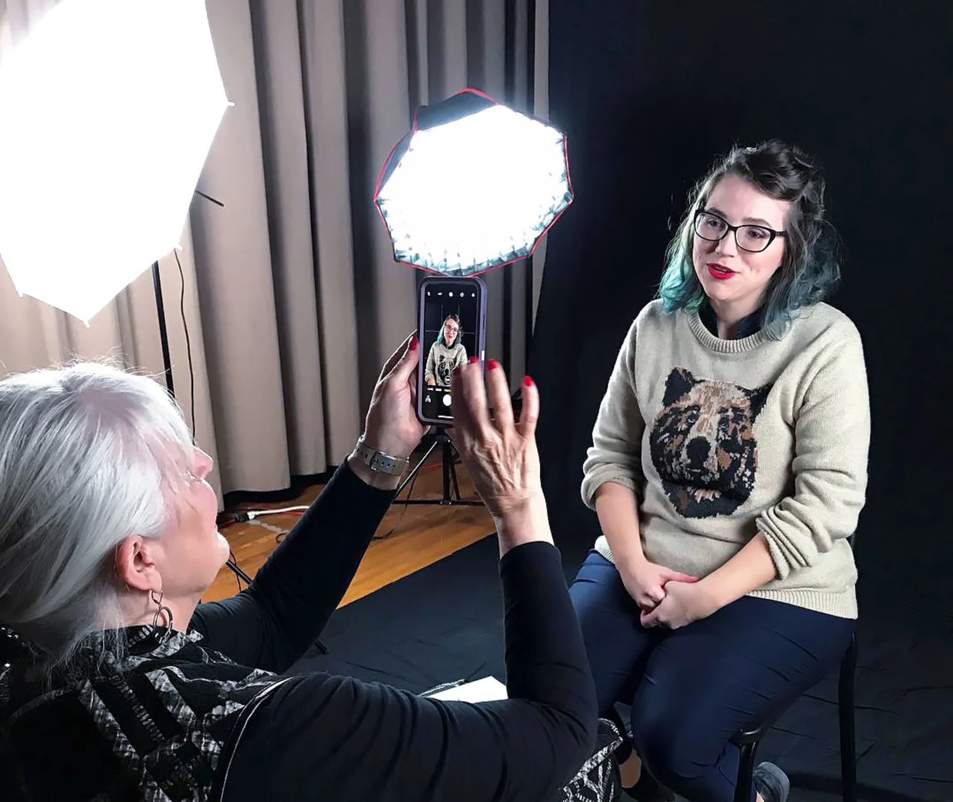 Beauty Thibodeau sits in a chair in front of a large black photography backdrop, lit from the side by several umbrella lights. She sits opposite Blessen, who is holding up an iPhone to take Thibodeau’s picture.