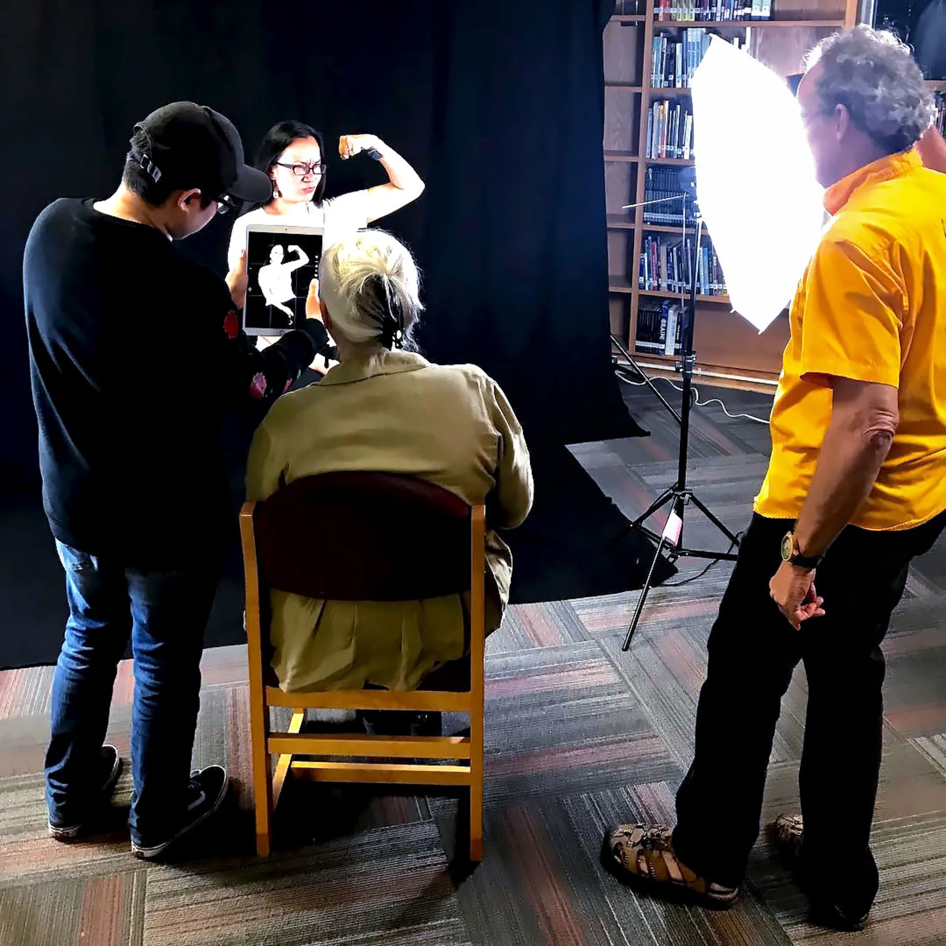 Wide shot of the photo shoot set, where several members of the 29 Pieces team adjust lighting and position an iPad to photograph Martinez, who is sitting in front of a black backdrop and flexing her bicep in a pose similar to Rosie the Riveter.