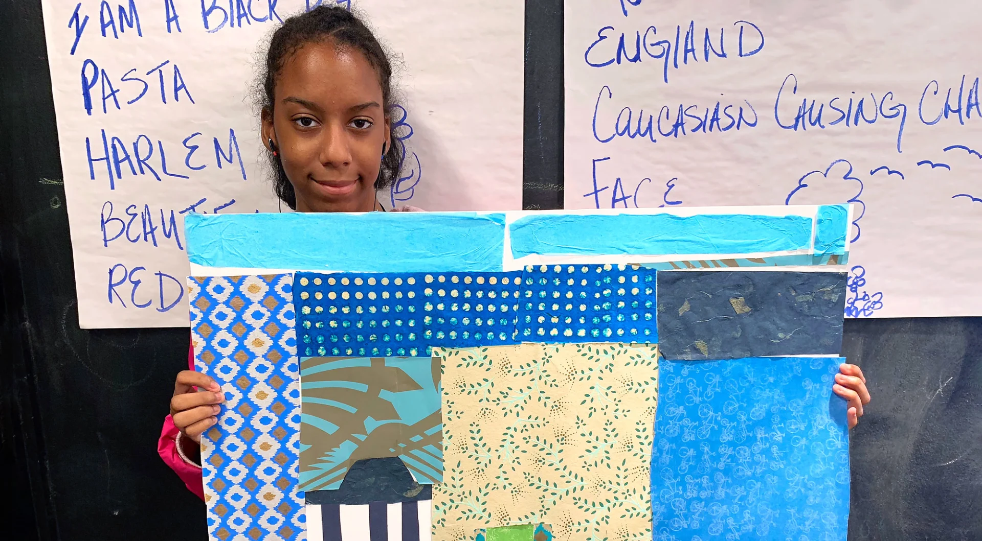 A young woman stands in front of a chalkboard with notes and holds up an abstract collage with multicolored patterned paper.