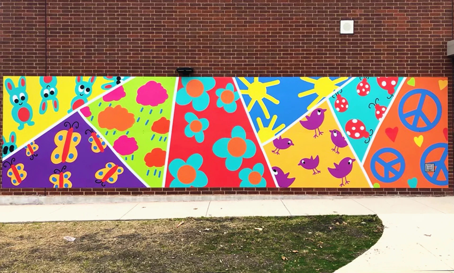 Rainbow-colored school mural against a brick wall, divided into sections featuring cartoon-style rabbits, butterflies, rainclouds, flowers, suns, birds, ladybugs, peace signs, and hearts.