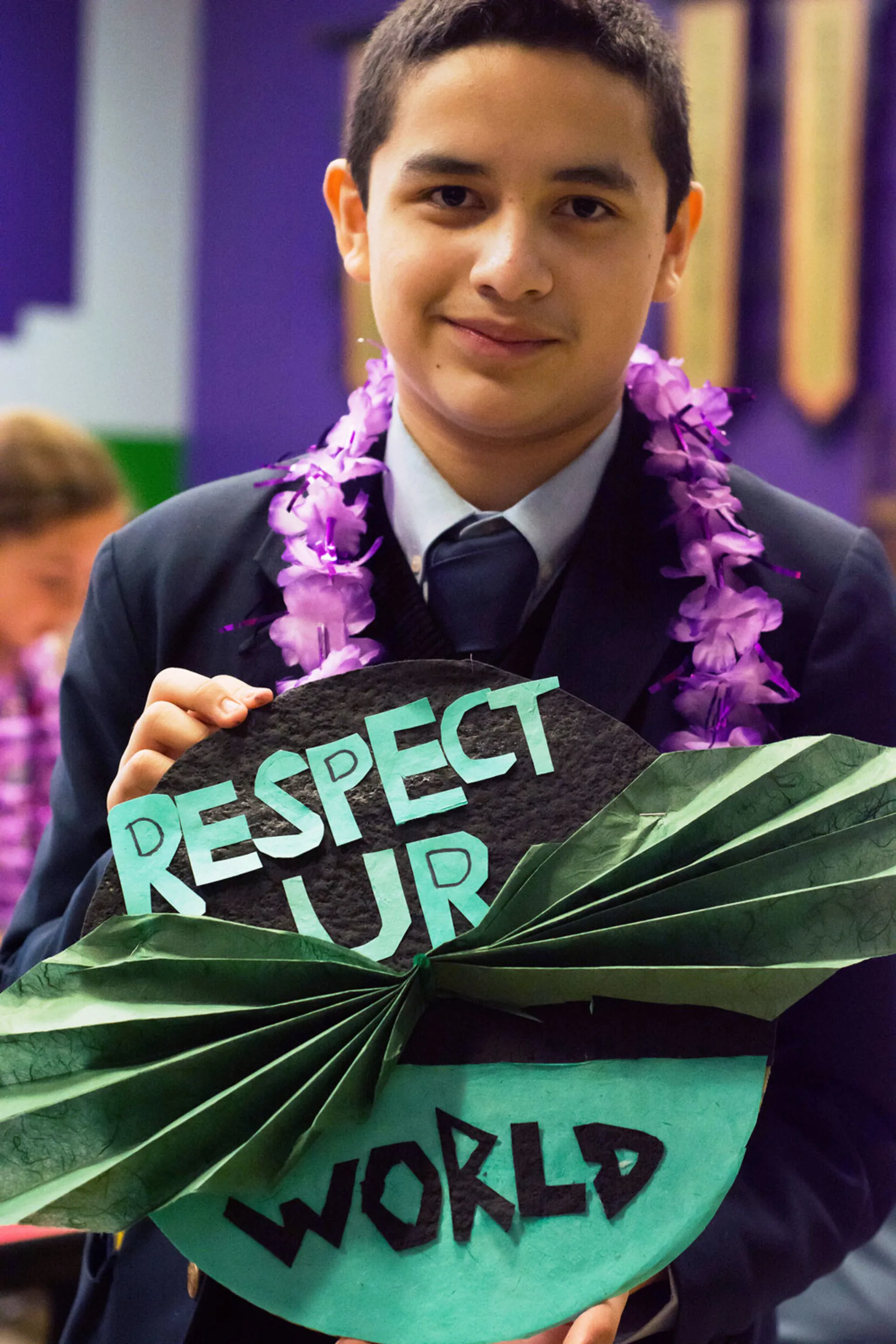 A teacher boy wearing a suit and a Hawaiian lei holds a collage decorated with folded green paper and the text, 'Respect Your [spelled UR] World'.