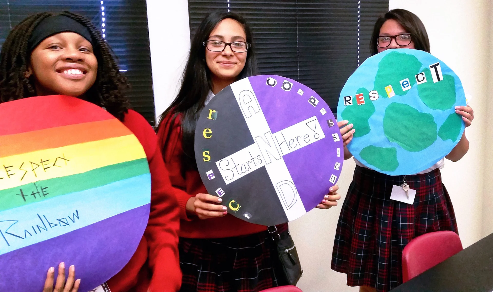 A group of 3 teenage girls smile and hold up circle-shaped respect artwork. One piece of art features a rainbow with the text 'Respect the Rainbow', while another includes the text, 'Respect and compassion starts here!', and a third shows planet Earth with the text, 'Respect'.