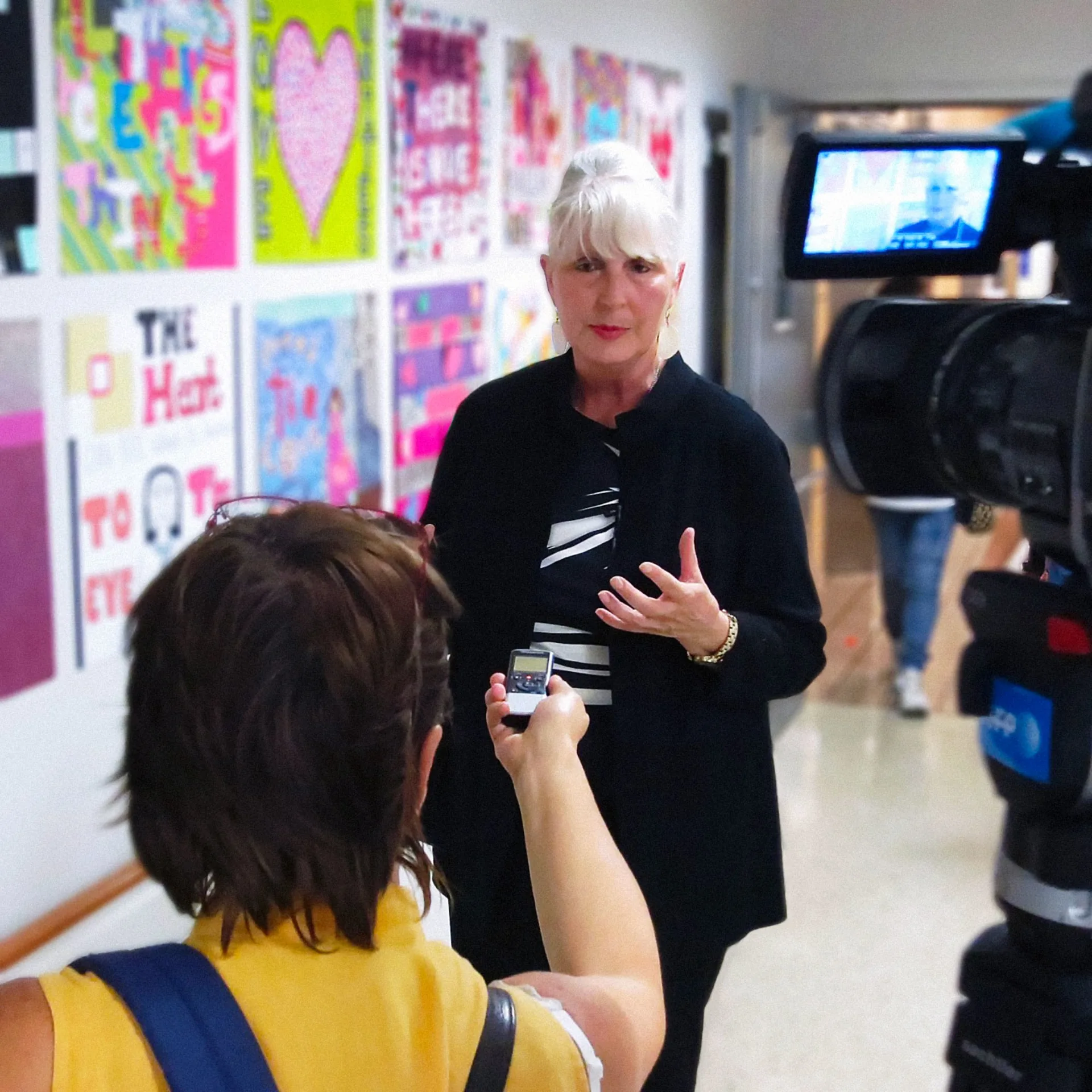 Blessen standing in a school hallway and speaking to a reporter with a microphone while a video crew records the interview.