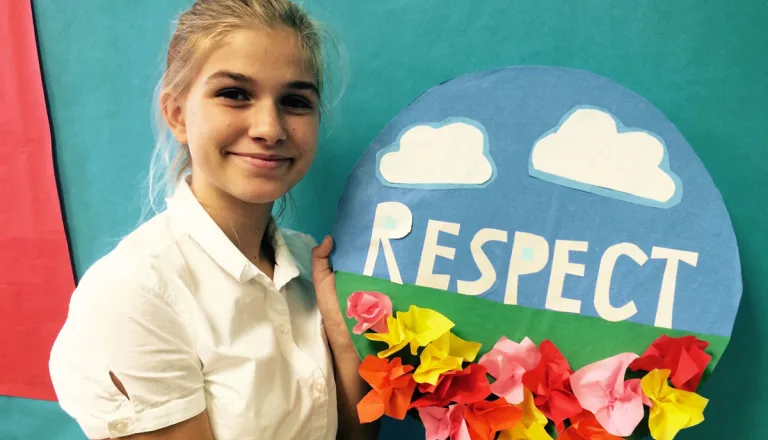A teenage girl holds up a circle-shaped collage with 3-dimensional red, pink and yellow paper flowers, clouds, and the word 'Respect'.