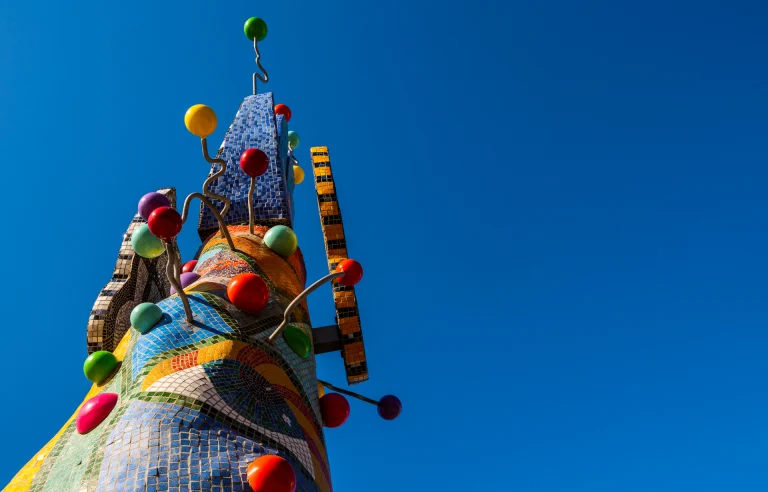 A low-angle view of the colorful, abstract Piece 24 sculpture set against a deep blue, cloudless sky. The base of the sculpture is adorned in bright, multicolored spheres and mosaic tiles that form patterns, including a large human eye.