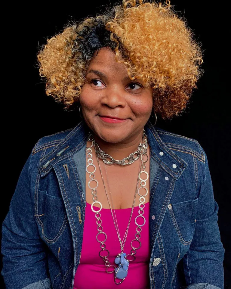 Dr. Smith, who is wearing a jean jacket and layers of necklaces, smiling and looking up to the right against a black photo backdrop.