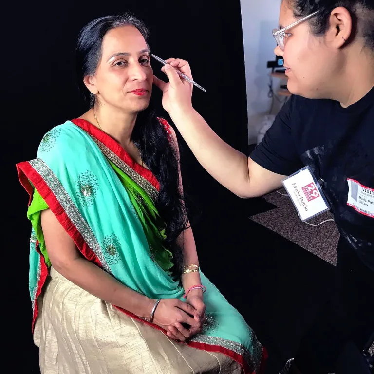 Chadha, who is wearing a multicolored sari, sits in a chair on the photo shoot set while Patiño uses a makeup brush to apply highlighter under her eyebrows.