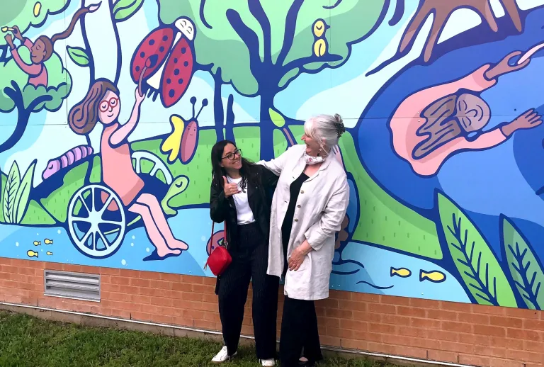 Alejandra Camargo and Karen Blessen hug and smile at each other while standing in front of the completed Burnet Elementary mural, which depicts children in a vibrant, whimsical nature scene.