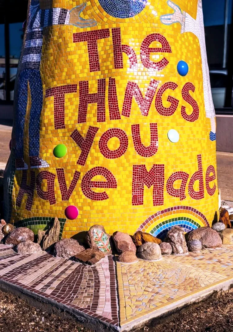 The base of Piece 24, which is decorated with mosaic tiles and the text, 'The things you have', resting on a flat, square display stand with rocks on a street in Dallas.