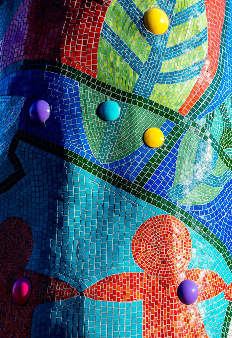 Close up of a section of Piece 24 featuring abstract mosaic shapes, leaves, and a chain of figures holding hands, along with shiny, multicolored half spheres.