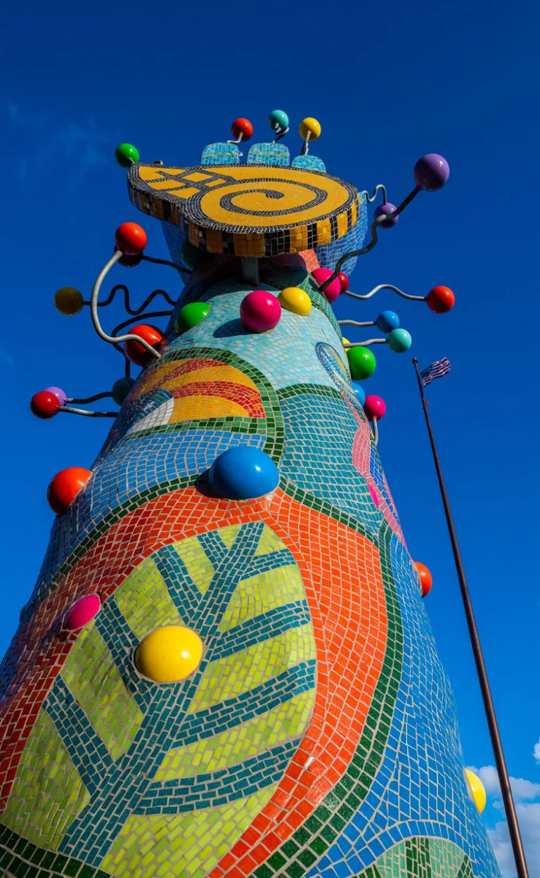 Low-angle view of the vibrant, multicolored Piece 24 against a deep blue sky. Red, pink, blue, yellow and green spheres protrude from the sculpture with wavy metal rods, and the top features a giant mosaic hand.