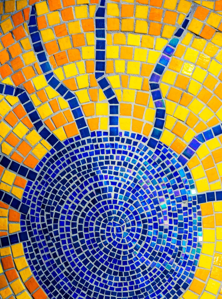 A section of Piece 24 featuring a vibrant sunburst design. The center of the sun is made up of concentric circles of small blue tiles, radiating outward into bright yellow and orange square tiles.