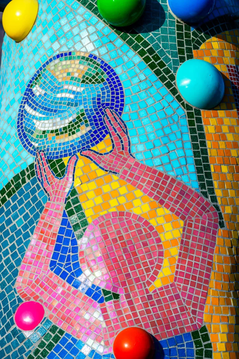 Colorful section of Piece 24 depicting a stylized human figure made of pink tiles, reaching up to hold a blue and green circular object resembling Earth. The background features vibrant patterns of blue, yellow, green, and orange tiles, with large, glossy, multicolored spheres embedded in the mosaic.