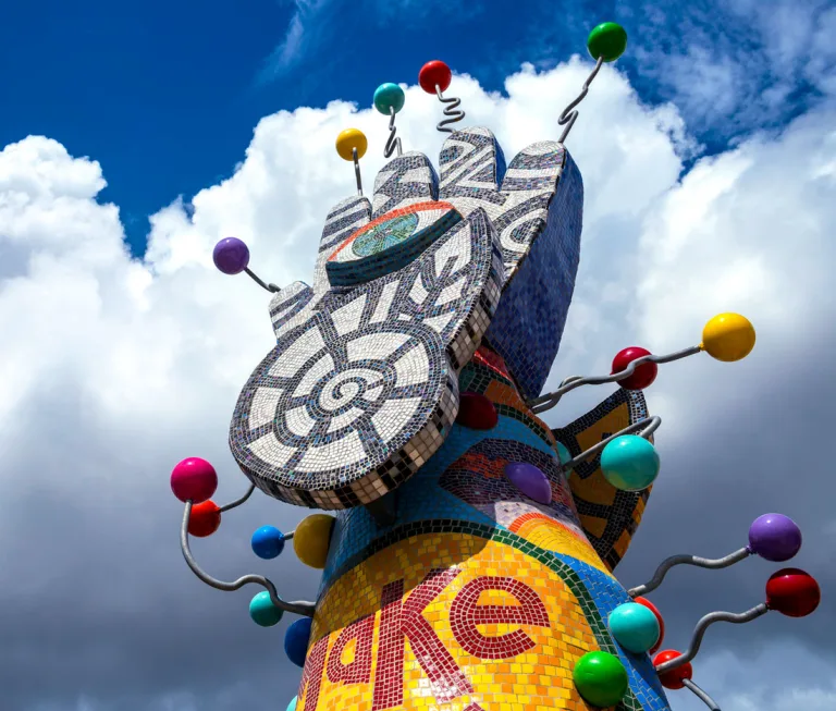 View of The top of Piece 24, which features a 3-dimensional mosaic hand with geometric patterns, an eye symbol in the center of the palm, and multicolored spheres extending from its fingertips with wavy metal rods.