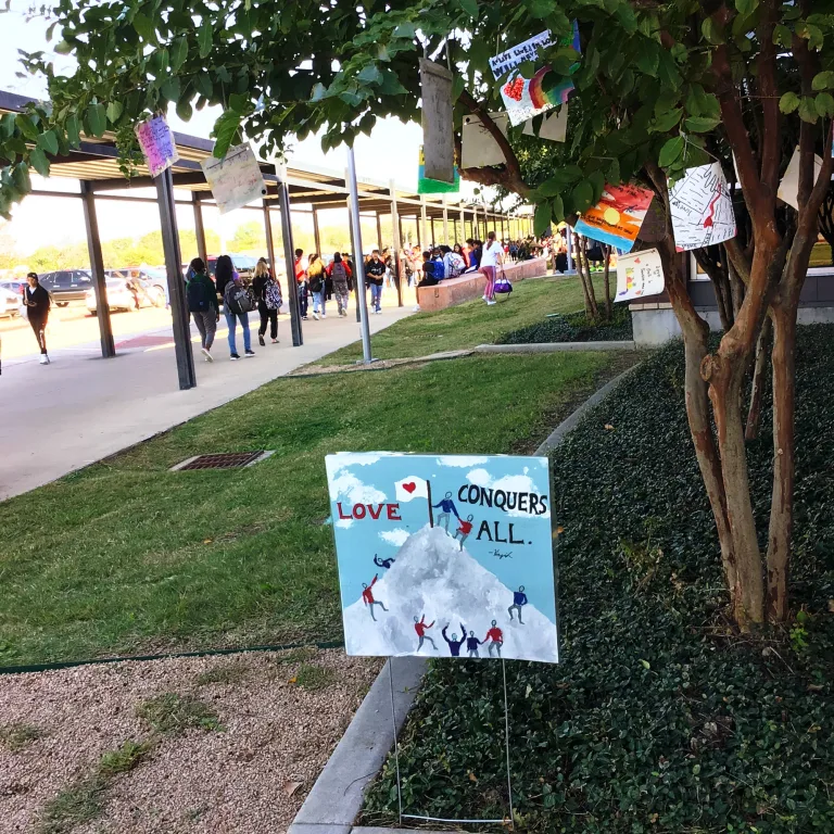 A yard sign in the grass beside a covered walkway with an illustration of people climbing a snowy mountain and planting a flag at the peak, along with text that reads 'Love Conquers All'.