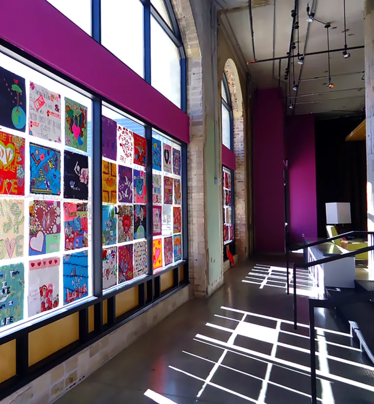 Sunlit school hallway with colorful, square love-themed artwork displayed in a grid inside window panels.