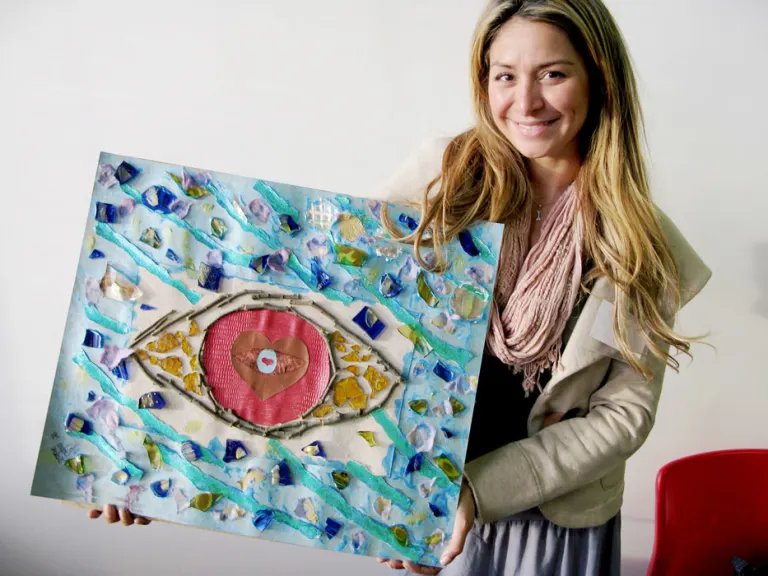 A woman smiles and holds up a collage of an eye surrounded by abstract shapes.