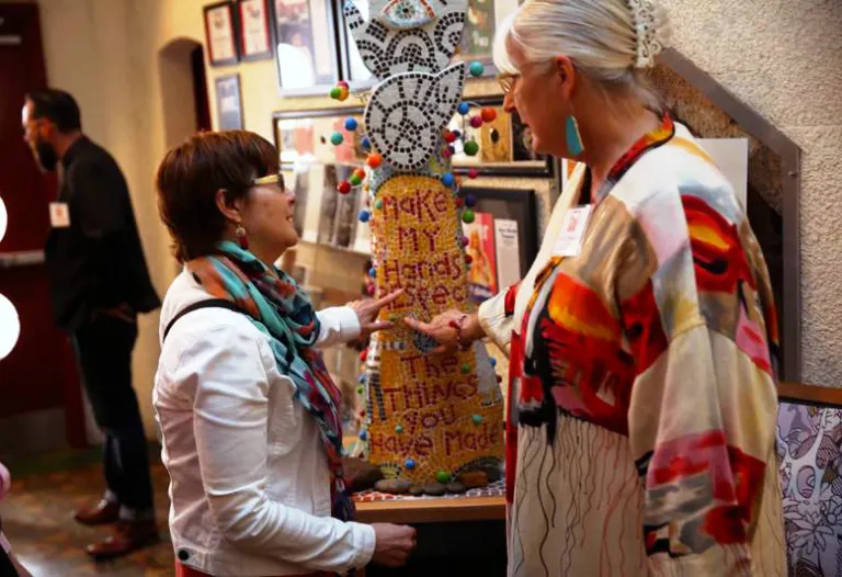 29 Pieces co-founder Karen Blessen and former Dallas Morning News Design Editor Marilyn Bishkin talking at a gathering while pointing to a prototype of the Piece 24 sculpture sitting on a table.